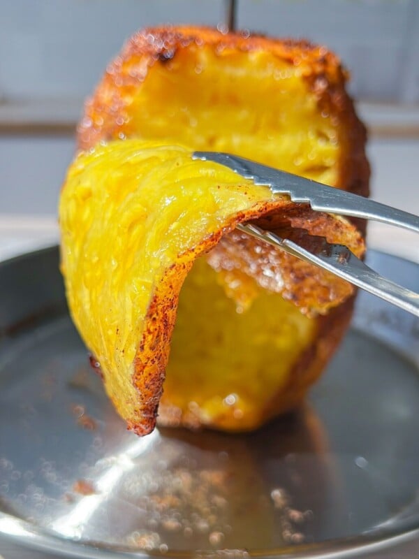 A pair of tongs lifts a crispy, golden-brown slice of Brazilian Grilled Pineapple from a larger piece on a skewer over a stainless steel tray.