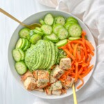 A bowl filled with sliced cucumbers, spiralized carrots, sliced avocado, and grilled chicken cubes—reminiscent of The Kourtney Kardashian Salad—all arranged neatly with two gold-colored forks on a white background.