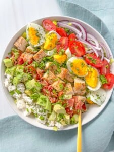 A colorful salad bowl featuring hard-boiled eggs reminiscent of classic egg sandwiches, grilled chicken, cherry tomatoes, red onion, avocado, bacon, cottage cheese, and herbs on fresh lettuce—served with a yellow fork.