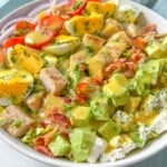 A bowl of chopped salad featuring grilled chicken, avocado, cherry tomatoes, hard-boiled eggs, bacon, red onion, lettuce, feta cheese, and a drizzle of yellow dressing—this classic Cobb Salad Recipe is both hearty and delicious.