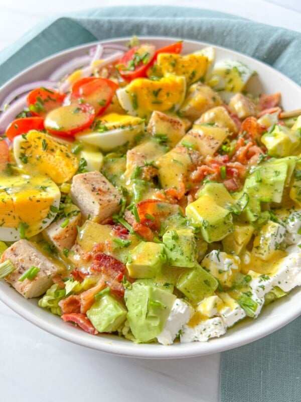 A bowl of chopped salad featuring grilled chicken, avocado, cherry tomatoes, hard-boiled eggs, bacon, red onion, lettuce, feta cheese, and a drizzle of yellow dressing—this classic Cobb Salad Recipe is both hearty and delicious.