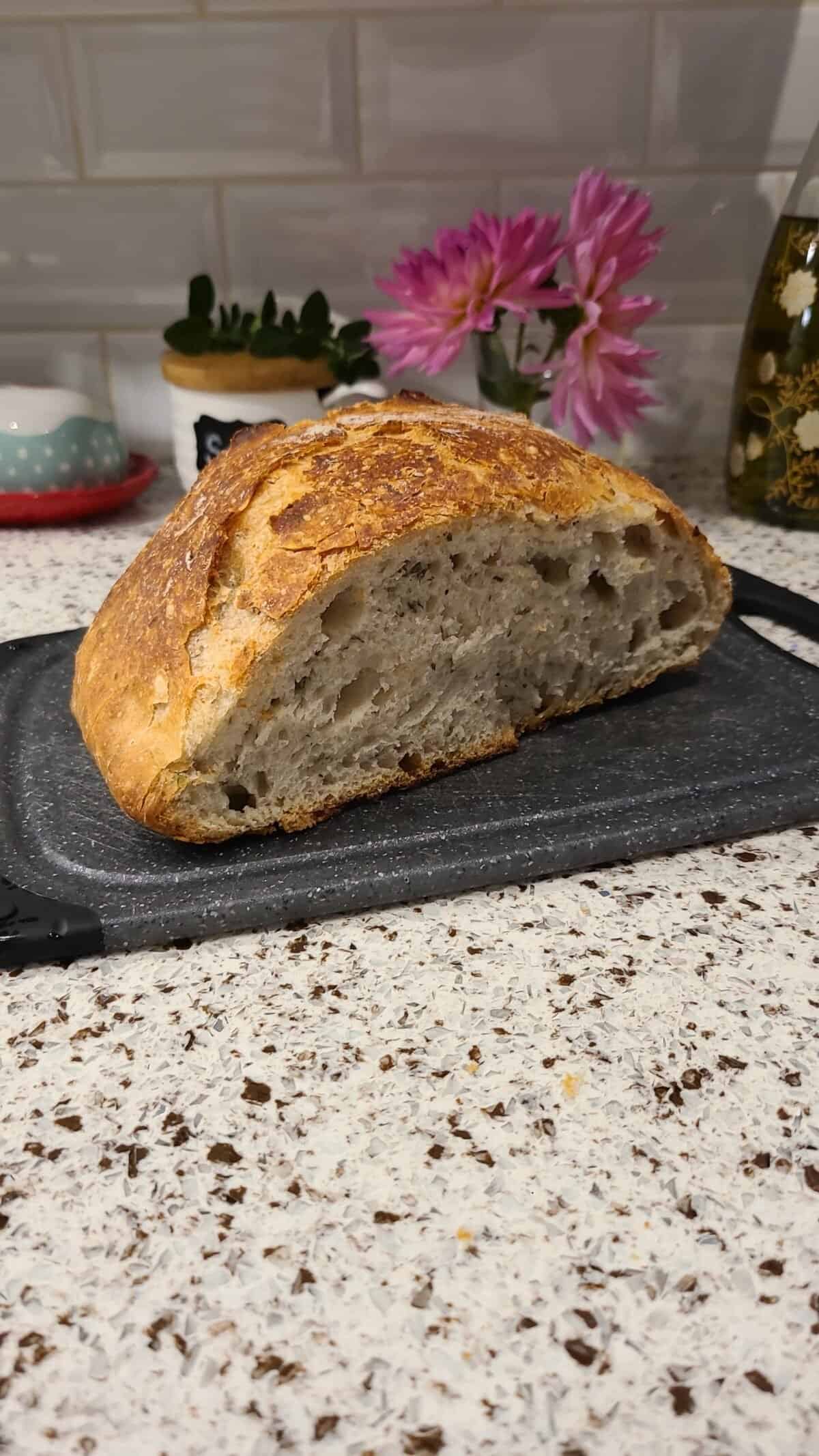 A loaf of no knead garlic bread with a crusty golden-brown exterior and airy interior sits sliced open on a cutting board. The background shows a speckled countertop, pink flowers, and kitchen items.