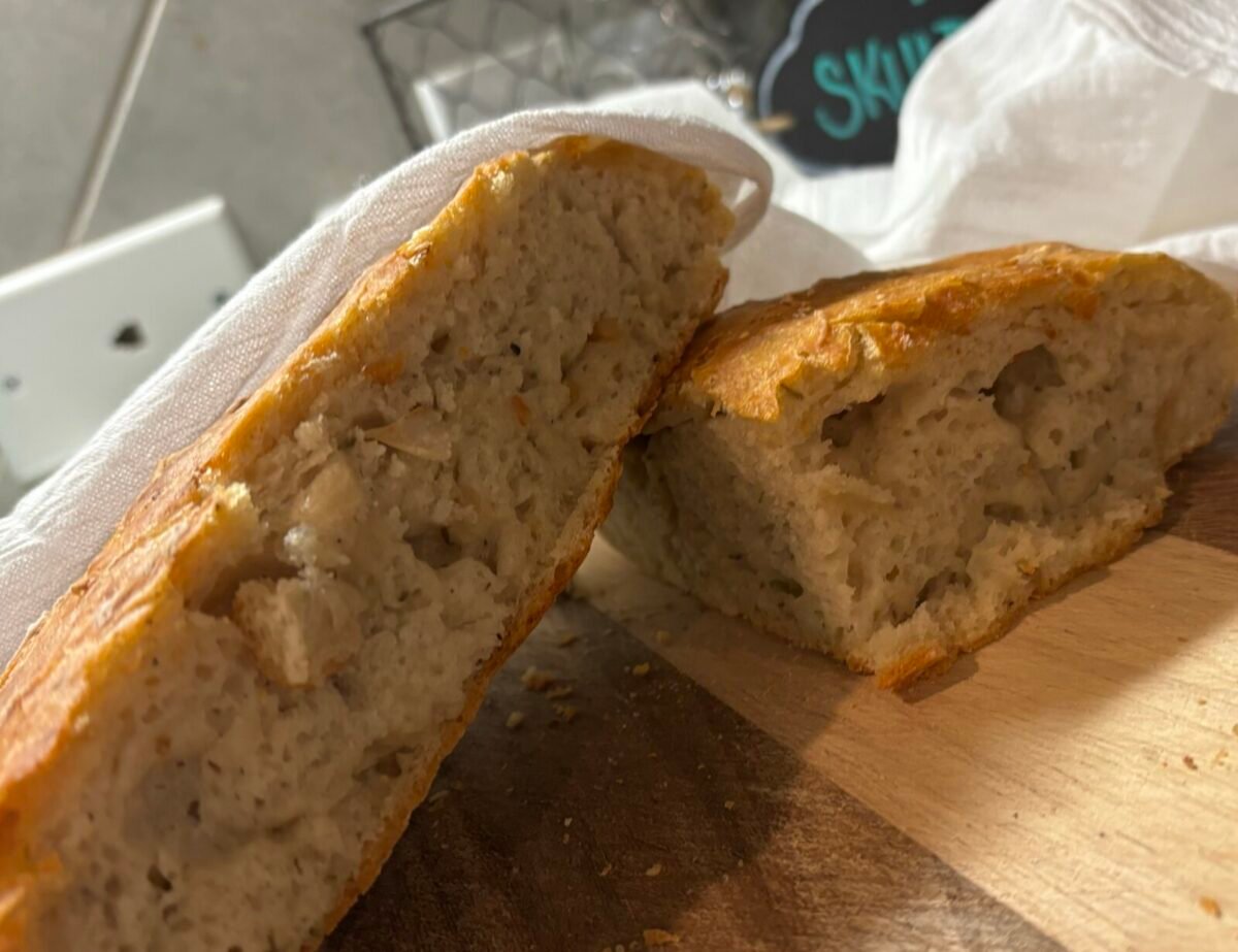 A loaf of rustic, No Knead Garlic Bread cut in half reveals a soft, airy interior. The bread sits on a wooden surface, partially covered by a white cloth, with crumbs scattered around the cutting board.