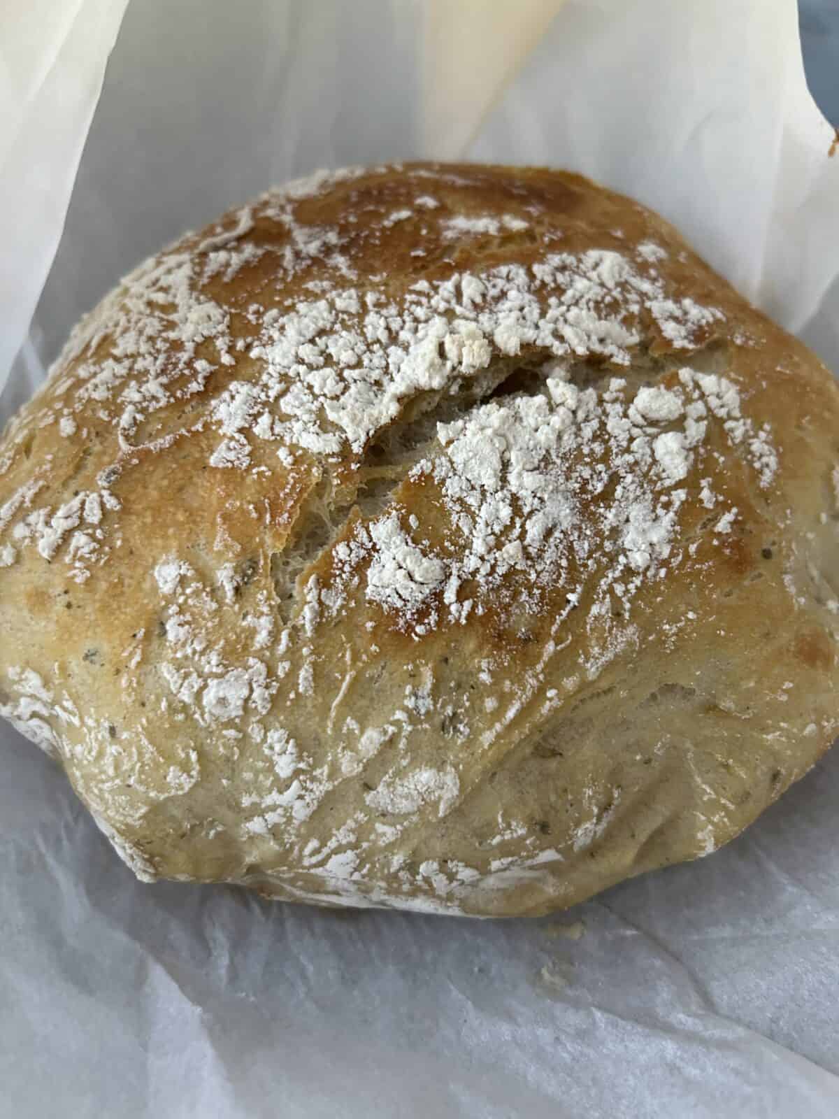 A round loaf of no knead garlic bread with a golden-brown crust, lightly dusted with white flour, rests on crumpled parchment paper.