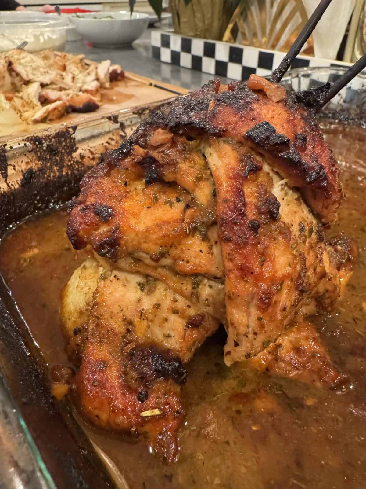 A roasted chicken in a baking dish, seasoned with herbs, sits in a pool of its own juices. Slices of grilled meat are in the background on a wooden cutting board, alongside a salad bowl and a checkered serving plate.