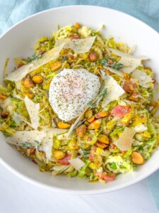 A bowl of shredded Brussels sprouts topped with a poached egg, shaved parmesan cheese, crispy bacon pieces, roasted pistachios, fresh herbs, and ground black pepper—just like The Kourtney Kardashian Salad.
