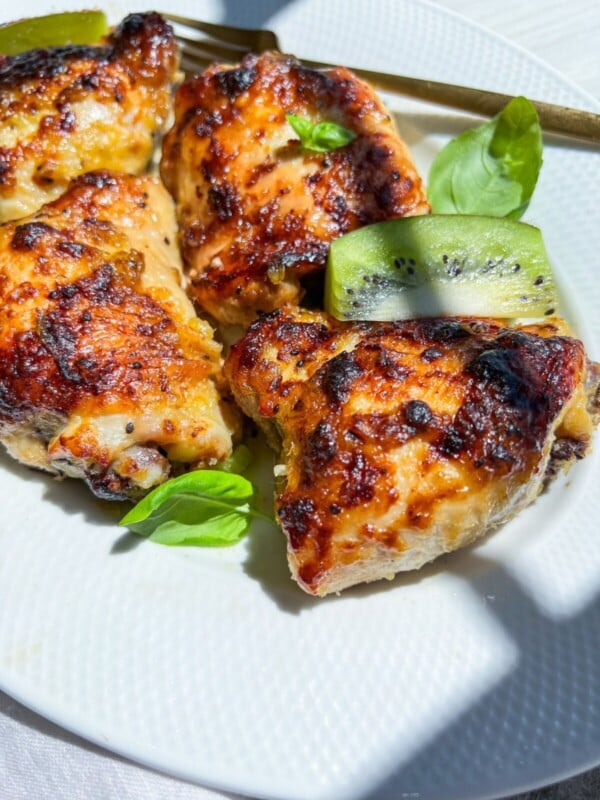 Three pieces of grilled, glazed chicken prepared with a zesty kiwi marinade for chicken, garnished with fresh basil leaves and a slice of kiwi, served on a white plate with a golden fork beside them.