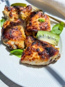 Close-up of three pieces of grilled, golden-brown chicken prepared with a zesty kiwi marinade for chicken, garnished with fresh basil leaves and slices of green kiwi on a white plate. Sunlight highlights the crispy texture.