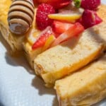 A close-up of fluffy, sliced French toast topped with fresh strawberries, raspberries, lemon slices, and drizzled with honey from a honey dipper, served on a white plate alongside warm No Knead Garlic Bread in bright natural light.
