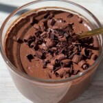 A glass bowl filled with creamy One Ingredient Chocolate Mousse, topped with chocolate shavings, and a spoon resting inside, all on a light wooden surface.