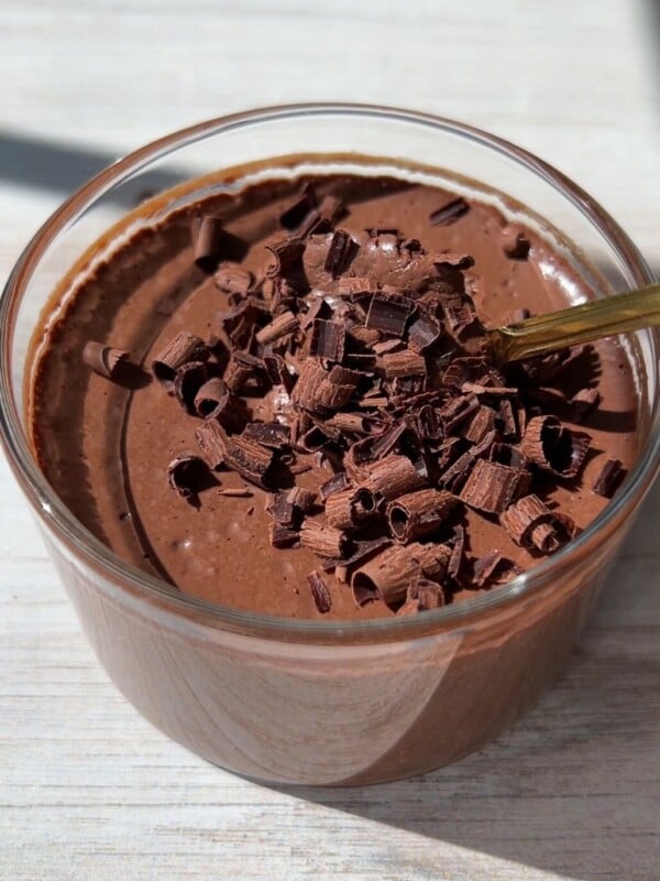 A glass bowl filled with creamy One Ingredient Chocolate Mousse, topped with chocolate shavings, and a spoon resting inside, all on a light wooden surface.
