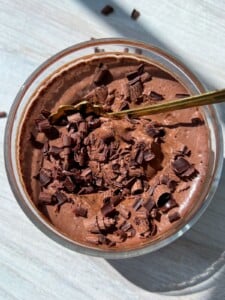 A glass bowl filled with creamy One Ingredient Chocolate Mousse, topped with dark chocolate shavings, and a gold spoon resting inside, all on a light-colored surface.