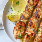 Two salmon skewers garnished with chopped herbs, served on a white plate with three lemon slices for garnish. The plate sits on a light-colored surface with a cloth partially visible.