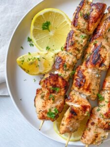 Two salmon skewers garnished with chopped herbs, served on a white plate with three lemon slices for garnish. The plate sits on a light-colored surface with a cloth partially visible.
