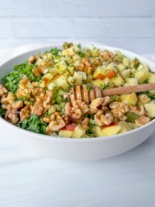 A white bowl filled with The Kourtney Kardashian Salad—chopped apples, walnuts, kale, and herbs—topped with a honey dipper and drizzled with golden honey.