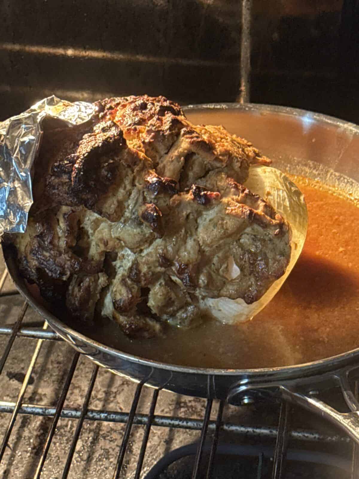 A large, browned roast sits in a pan filled with juices inside an oven. Part of the roast is covered with foil, and the surface is crispy and golden from roasting—reminiscent of perfectly cooked Greek Chicken Gyros.