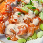 A close-up of a Spicy Salmon Poke Bowl featuring diced raw salmon, edamame beans, sesame seeds, chopped green onions, and creamy dressing, served on a white plate with chopsticks.