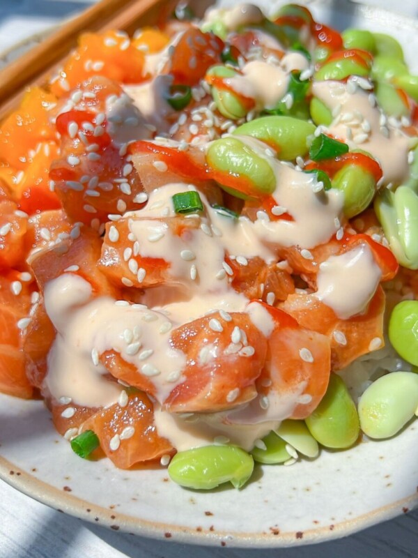 A close-up of a Spicy Salmon Poke Bowl featuring diced raw salmon, edamame beans, sesame seeds, chopped green onions, and creamy dressing, served on a white plate with chopsticks.
