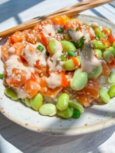 A Salmon Rice Bowl featuring diced salmon, edamame, rice, green onions, sesame seeds, and creamy sauce is served with chopsticks on a white plate in natural sunlight.