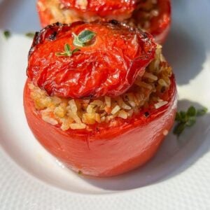 A close-up of a baked tomato stuffed with rice and herbs, topped with a roasted tomato slice and garnished with fresh thyme, served on a white plate.
