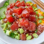 A Tuna Poke Bowl featuring diced raw tuna, edamame, sliced cucumber, chopped green onions, sesame seeds, and chunks of orange fruit, served in a white bowl with wooden chopsticks.