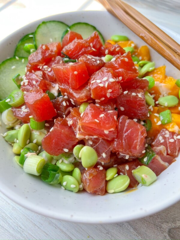 A Tuna Poke Bowl featuring diced raw tuna, edamame, sliced cucumber, chopped green onions, sesame seeds, and chunks of orange fruit, served in a white bowl with wooden chopsticks.