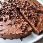 A round Two Ingredient Cake topped with chocolate shavings sits on a white plate. A single slice has been cut and slightly pulled out, revealing the smooth, rich chocolate interior.