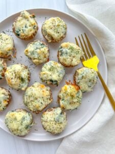 A plate of Spinach Stuffed Mushrooms topped with melted cheese and herbs, arranged neatly next to a yellow fork on a light-colored tablecloth.