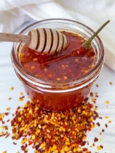 A glass jar filled with chili honey, topped with a whole chili pepper and a honey dipper. Crushed red pepper flakes are scattered around the jar on a white surface—perfect inspiration for your next Hot Honey Recipe.