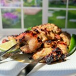 Three metal skewers with grilled chicken kebabs, inspired by Whole Lemon Chicken flavors, are served on a white plate, garnished with a lime wedge and a green chili. Sunlight streams in from a window, revealing a garden view in the background.
