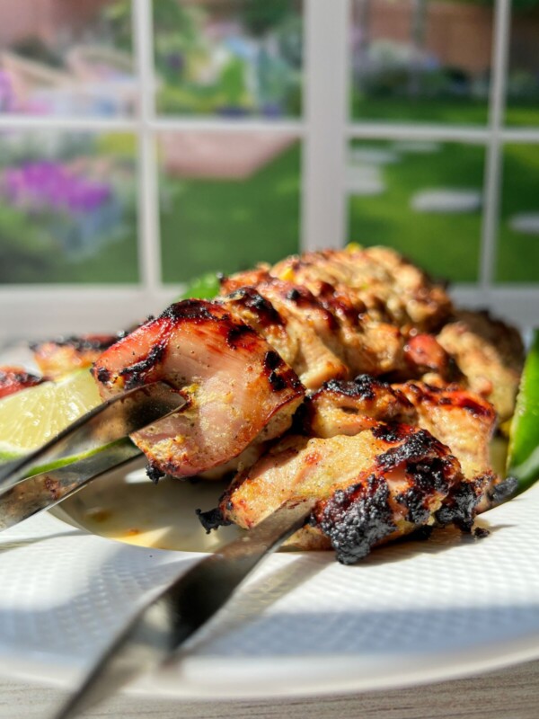 Three metal skewers with grilled chicken kebabs, inspired by Whole Lemon Chicken flavors, are served on a white plate, garnished with a lime wedge and a green chili. Sunlight streams in from a window, revealing a garden view in the background.