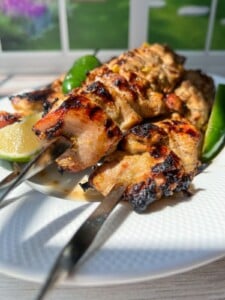 Grilled chicken skewers on a white plate, garnished with green chilies and a slice of lime, evoke the zesty flavors of Whole Lemon Chicken, with a bright window and garden view in the background.