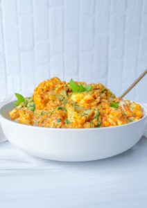 A white bowl filled with creamy cauliflower pasta mixed with vegetables and herbs sits on a white surface, with a gold fork partially visible inside the bowl. The background is a light-colored, textured wall.
