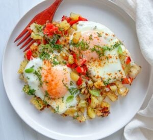 Two fried eggs topped with chopped herbs sit on a bed of diced potatoes and red bell peppers, creating a classic breakfast skillet on a white plate, with a wooden fork and white napkin beside the dish.