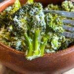 A close-up of Creamy Baked Broccoli florets coated in a luscious white sauce, with a fork lifting one piece above a wooden bowl brimming with more broccoli.