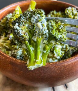 A close-up of Creamy Baked Broccoli florets coated in a luscious white sauce, with a fork lifting one piece above a wooden bowl brimming with more broccoli.