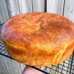 A large, round loaf of Portuguese Sweet Bread with a golden-brown crust is cooling on a black wire rack, with a wooden fence in the background.