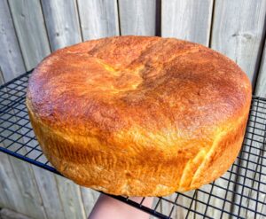 A large, round loaf of Portuguese Sweet Bread with a golden-brown crust is cooling on a black wire rack, with a wooden fence in the background.