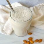 A glass of frothy almond milk made from a homemade almond milk recipe sits on a white surface, surrounded by whole almonds and a cream-colored cloth in the background.