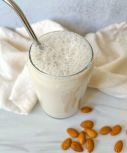 A glass of frothy almond milk made from a homemade almond milk recipe sits on a white surface, surrounded by whole almonds and a cream-colored cloth in the background.