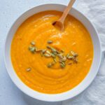 A bowl of creamy Roasted Butternut Squash Soup topped with pumpkin seeds, with a wooden spoon resting inside. The bowl sits on a light surface next to a textured white cloth.