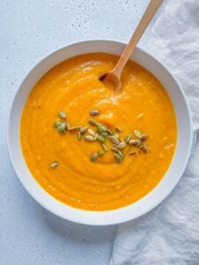 A bowl of creamy Roasted Butternut Squash Soup topped with pumpkin seeds, with a wooden spoon resting inside. The bowl sits on a light surface next to a textured white cloth.