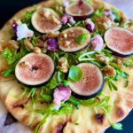 A flavorful Naan Pizza topped with fresh arugula, sliced figs, walnuts, goat cheese crumbles, and a drizzle of dressing served on a wooden board.