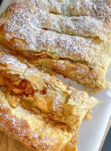 A close-up of a sliced, golden-brown puff pastry strudel with flaky layers, filled with apple pieces and dusted with powdered sugar, resting on parchment paper.