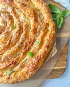 A golden, spiral-shaped pastry rests on parchment paper atop a wooden board, garnished with a few fresh green leaves. The pastry has a flaky, crisp texture and appears freshly baked.