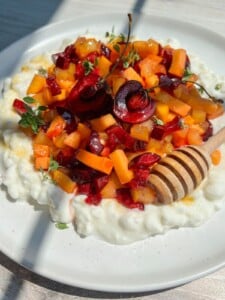 A plate of creamy Cottage Cheese Fruit Salad topped with chopped peaches, cherries, fresh thyme, and a honey dipper drizzling honey, all on a white plate with sunlight casting soft shadows.