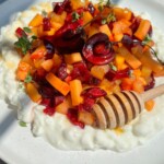 A plate of creamy Cottage Cheese Fruit Salad topped with chopped cherries, apricots, fresh herbs, and a honey dipper drizzled with honey, served on a white dish in bright natural light.