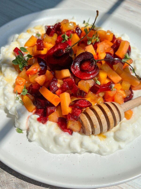 A plate of creamy Cottage Cheese Fruit Salad topped with chopped cherries, apricots, fresh herbs, and a honey dipper drizzled with honey, served on a white dish in bright natural light.