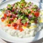 A vibrant Cottage Cheese Salad featuring diced cucumbers, tomatoes, red onions, and herbs drizzled with olive oil comes together as a refreshing chopped salad on a white plate bathed in sunlight.