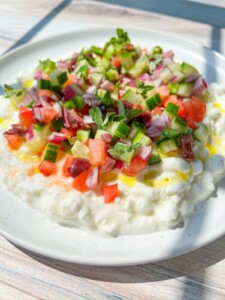A vibrant Cottage Cheese Salad featuring diced cucumbers, tomatoes, red onions, and herbs drizzled with olive oil comes together as a refreshing chopped salad on a white plate bathed in sunlight.